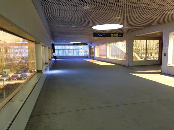 Photo from inside the bridge over the railway that passes through the center of Lund; the yellow filters on the windows make white light from the outside world look a bright, almost Cherenkov radiation blue, and create an orange patch of sunlight on the concrete floor, in this liminal space with a metal mesh ceiling. A sign points towards the steps for ”Spår 2-3”.