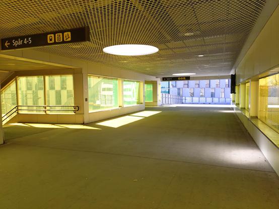 Photo from inside the bridge over the railway that passes through the center of Lund, in the other direction. The yellow and lime-green filters on the windows make the white light from the outside world look a bright, almost lavender blue, and create bright green and white patches of sunlight on the concrete floor, in this liminal space with a metal mesh ceiling. A sign points towards the steps for ”Spår 4-5”. In the distance, in the blue, a winding ramp for bikes and pedestrians curves off into the distance, with metal railings, and behind it a very modern building with alternating glass and concrete panels in a checkerboard pattern.